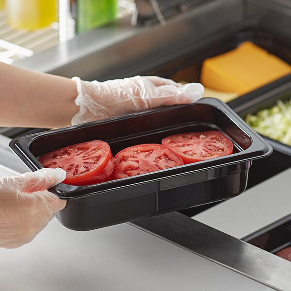 A person in gloves holding a Vigor black polycarbonate food pan with tomatoes.