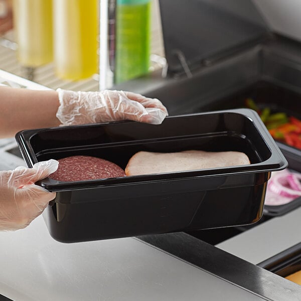 A person in gloves holding a Vigor black polycarbonate food pan full of meat.