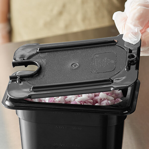A gloved hand placing a black Vigor polycarbonate food pan lid on a plastic container of onions.