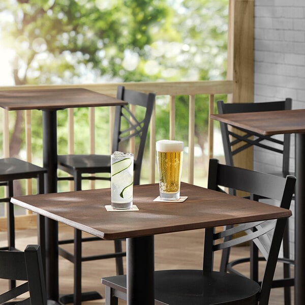 A Lancaster Table & Seating Excalibur square table with a textured walnut finish and a glass of beer on it.