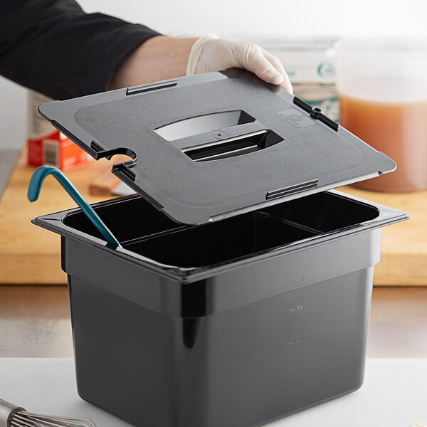 A hand holding a black Vigor 1/2 size plastic food pan lid with a notch and handle over a black container.