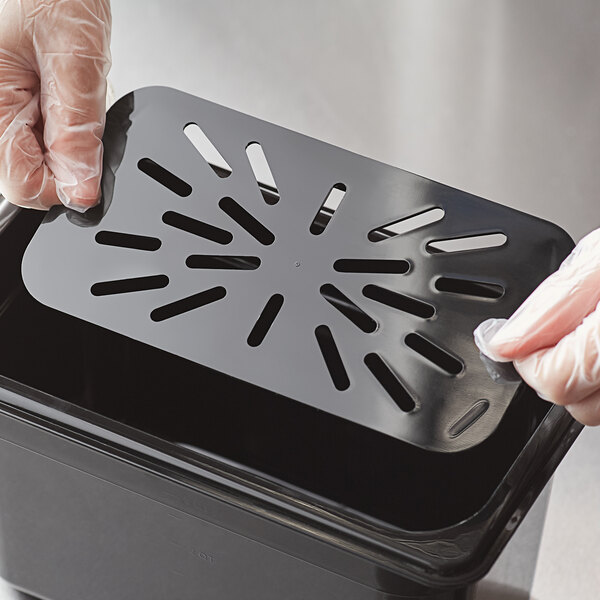 A person in gloves cleaning a black Vigor 1/4 size polycarbonate drain tray over a black trash can.