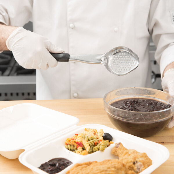 A person in a white chef's coat using a Vollrath black perforated oval Spoodle over a bowl of food.