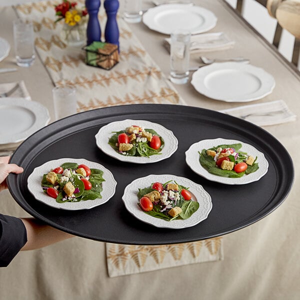 A person holding a Choice black oval non-skid serving tray with four small plates of food on it.