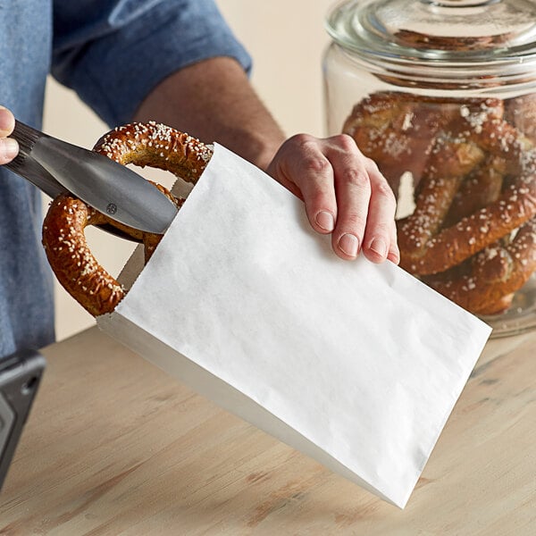A hand using a knife to cut a pretzel inside a white paper bag.