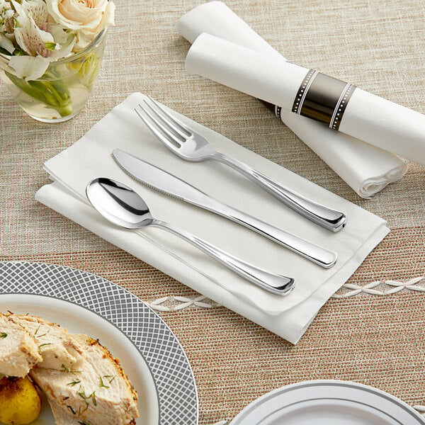 A table setting with a Visions white linen-feel napkin and classic silver plastic cutlery on a table.