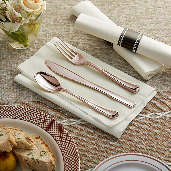 A Visions white pre-rolled napkin with rose gold plastic cutlery on a table with a plate of food and a glass with flowers.