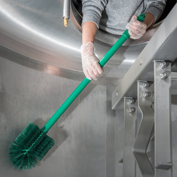 A person in gloves using a Carlisle green brush with a green handle to clean a metal container.