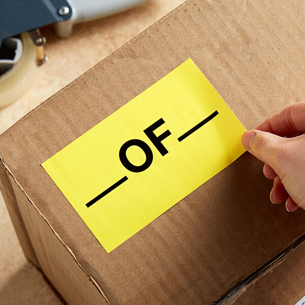A hand using a Lavex matte paper label to apply a yellow sign to a cardboard box.