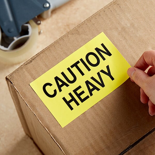 A hand using a Lavex Caution Heavy Matte Paper Label to put a yellow caution sticker on a cardboard box.