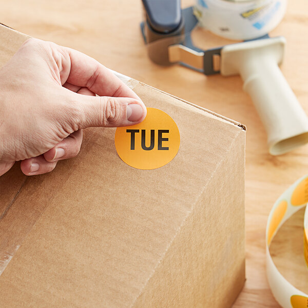 A hand holding a Lavex orange matte paper sticker and applying it to a cardboard box.
