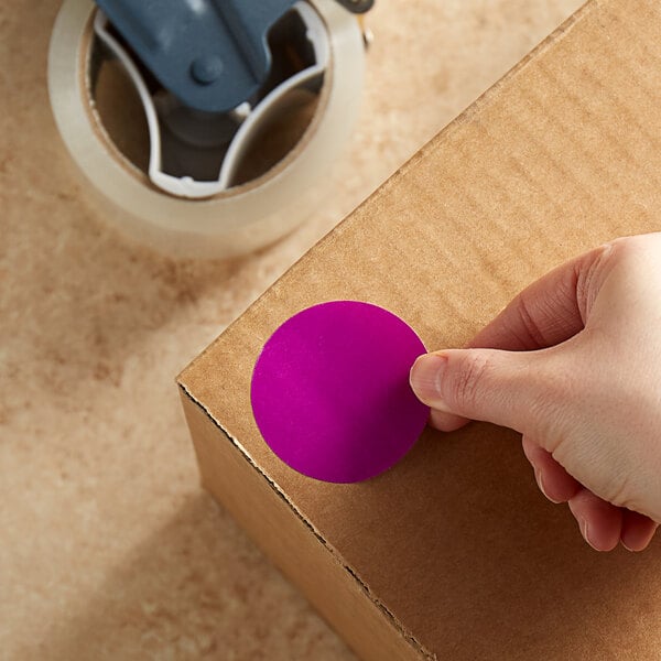 A hand holding a Lavex purple round inventory label on a cardboard box.