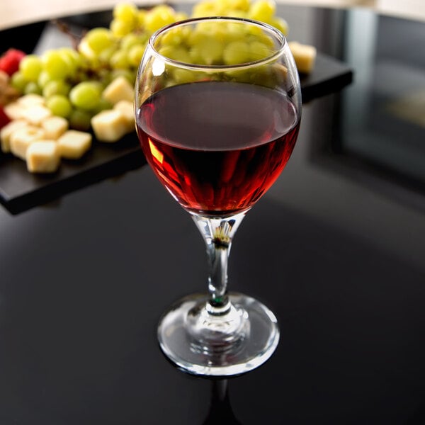 A Libbey wine glass filled with red wine and grapes on a table.