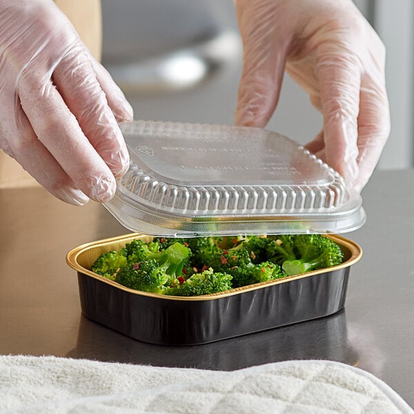 A hand in plastic gloves placing a dome lid on a ChoiceHD black and gold mini foil container filled with broccoli.