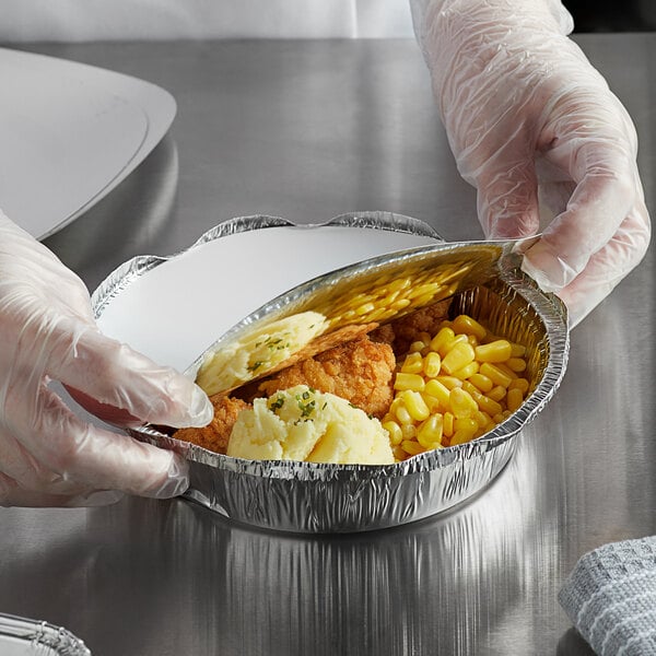 A person in gloves holding a Choice foil-laminated board lid over a tray of food.