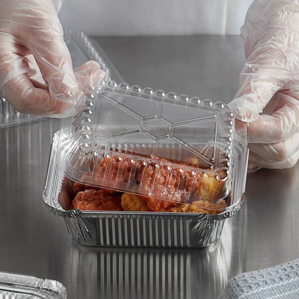 A hand wearing a plastic glove holding a Choice clear dome lid on a container of chicken.