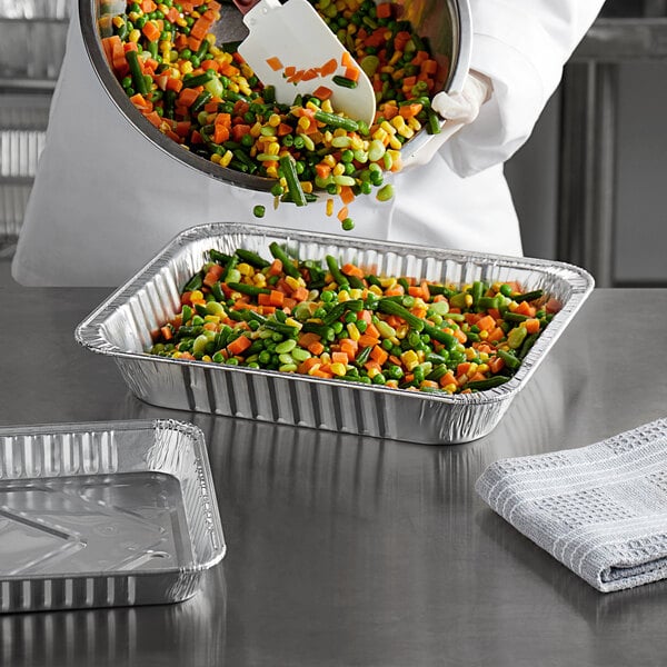 A chef pouring mixed vegetables into a Choice half size heavy-duty foil steam table pan.