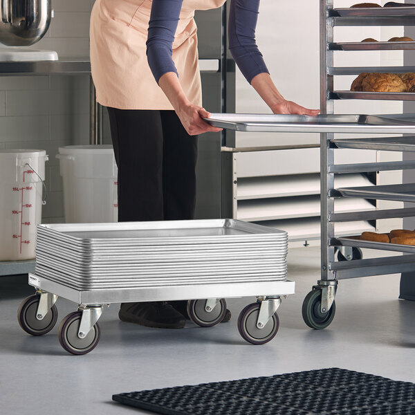 A stack of metal trays is placed on a low, wheeled dolly in a commercial kitchen setting, with a person transferring a tray to a nearby rack.