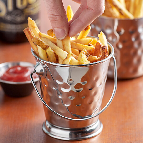 A person holding a Vollrath mini stainless steel serving bucket filled with french fries dipped in ketchup.