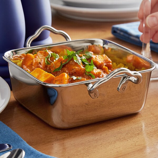 A person using a fork and knife to eat food from a Vollrath stainless steel mini roasting pan with handles.