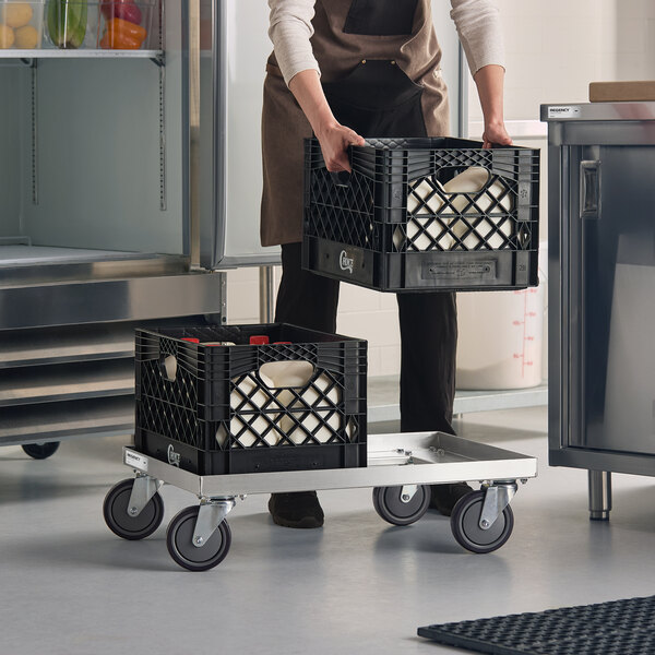 A person is placing a black plastic crate filled with jugs onto a metal dolly with four wheels, next to another similar crate already on the dolly.