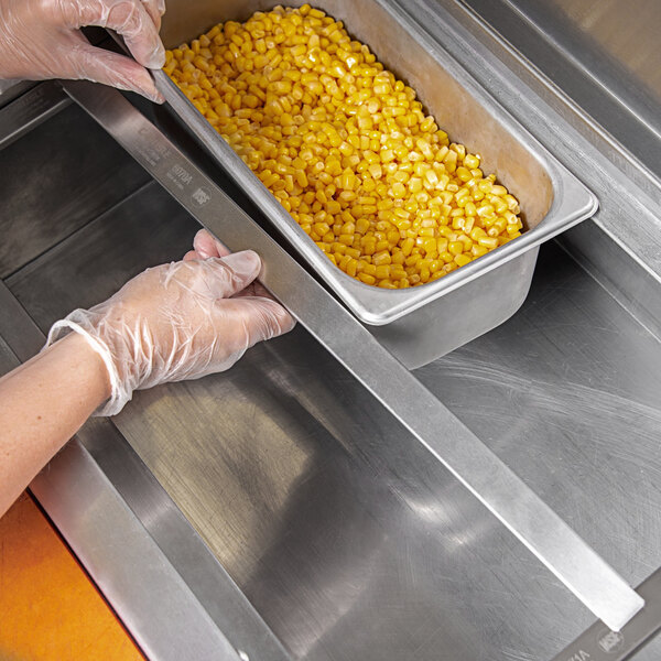 A gloved hand using a Carlisle stainless steel steam table pan adapter to hold a container of corn.