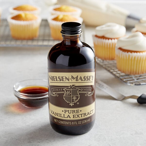 A bottle of Nielsen-Massey Pure Vanilla Extract next to cupcakes on a table.