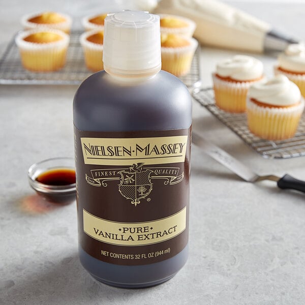 A bottle of Nielsen-Massey Pure Vanilla Extract on a table next to a cupcake with white frosting.