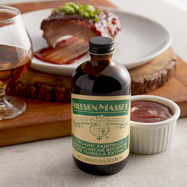 A black bottle of Nielsen-Massey Organic Madagascar Bourbon Vanilla Extract on a table next to a plate of ribs.