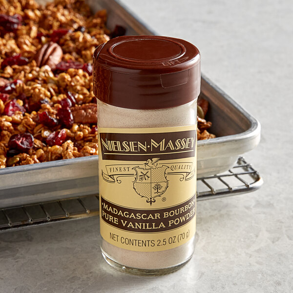 A close-up of a jar of Nielsen-Massey Madagascar Bourbon Pure Vanilla Powder on a counter next to a tray of granola.
