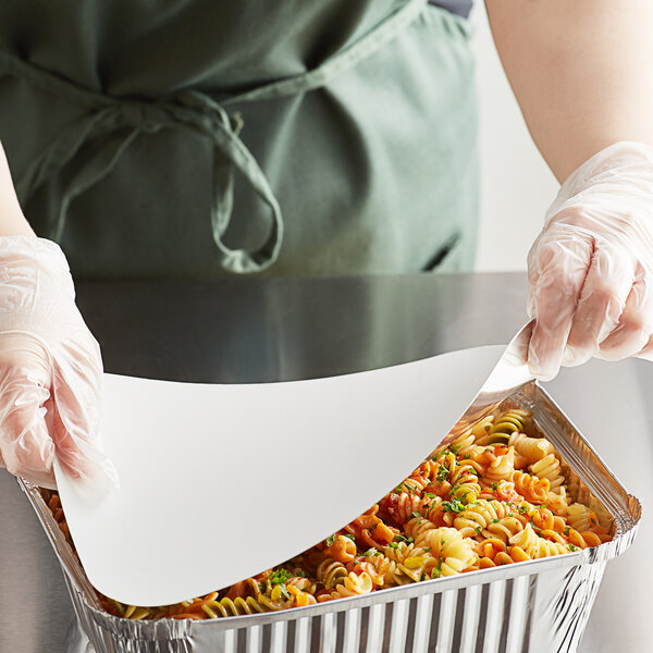 A person in gloves holding a Choice foil-laminated board lid over a tray of pasta.
