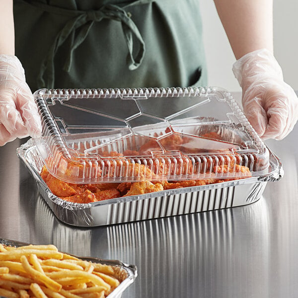 A person in gloves holding a tray of food with a Choice oblong foil container and dome lid.