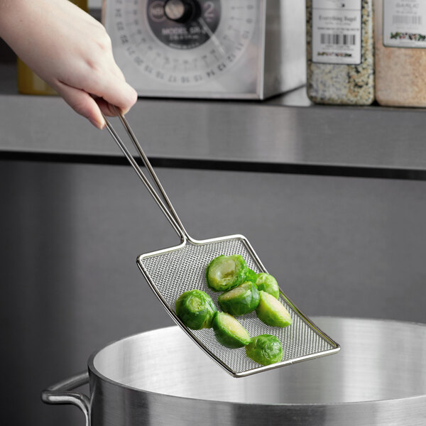 A hand holding a Tablecraft nickel mesh skimmer with brussels sprouts over a pot.
