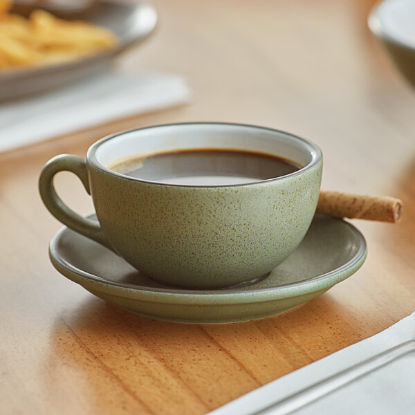 An Acopa Embers moss green stoneware cup of coffee on a saucer on a wooden table.