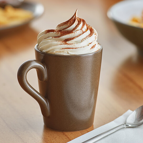 A close-up of a Acopa Embers hickory brown stoneware mug filled with hot chocolate and whipped cream with a spoon.