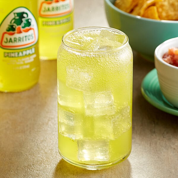 A close-up of a glass of yellow Jarritos pineapple soda with ice on a table with chips.