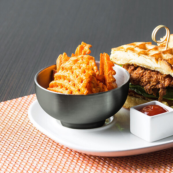 A plate with a sandwich and fries with a matte black stainless steel bowl of red sauce.