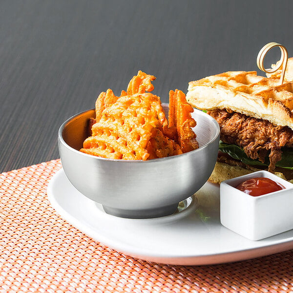 A plate with a sandwich and fries and a stainless steel bowl of red sauce on a table.
