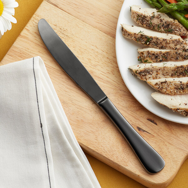 An Acopa Vernon stainless steel dinner knife on a cutting board next to a plate of chicken and asparagus.