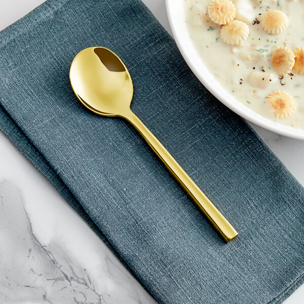 An Acopa Phoenix stainless steel bouillon spoon with a gold handle on a blue napkin next to a bowl of soup.