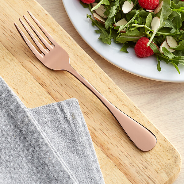 An Acopa Vernon Rose Gold stainless steel salad fork on a wooden board next to a plate of salad.