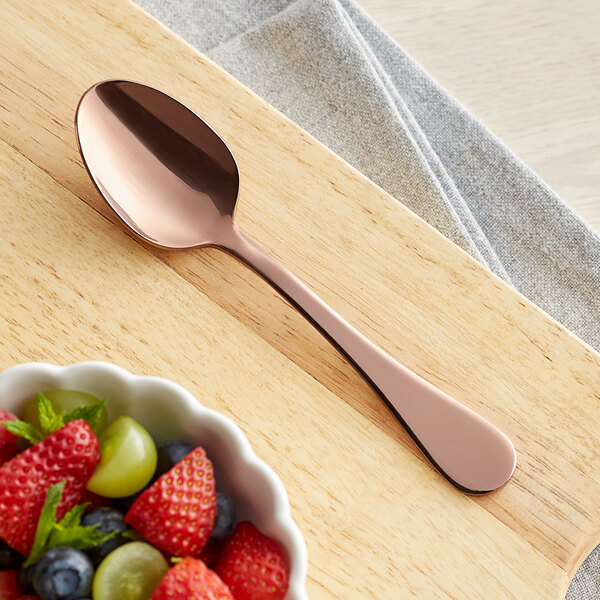 An Acopa Vernon rose gold stainless steel teaspoon on a table with a bowl of fruit.
