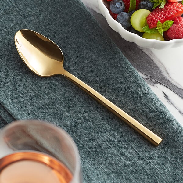 An Acopa Phoenix stainless steel teaspoon with a gold handle on a cloth next to a bowl of fruit.