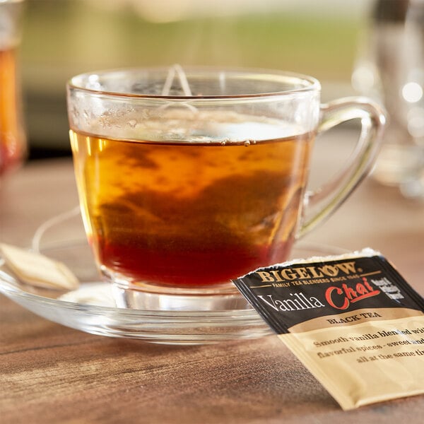 A glass cup of Bigelow Vanilla Chai tea with a tea bag on a saucer.