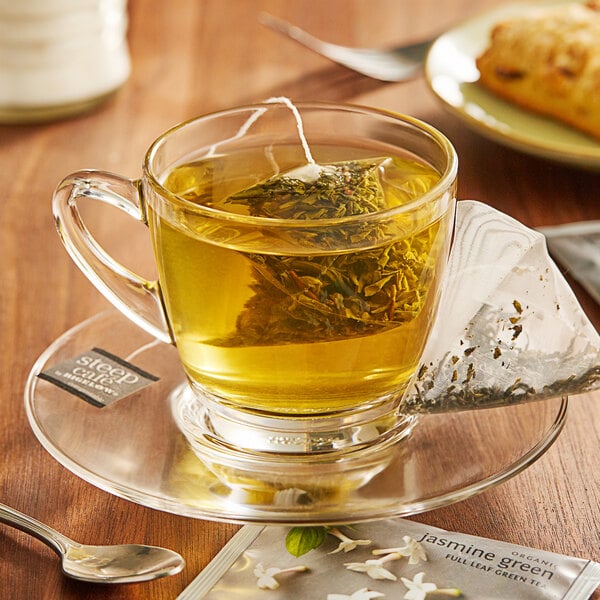 A glass cup of Steep Cafe By Bigelow Organic Jasmine Green Tea with a bag of tea on a saucer.