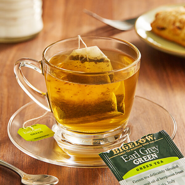 A glass cup of Bigelow Earl Grey green tea with a tea bag on a saucer.