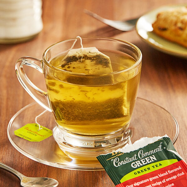 A glass cup of Bigelow Constant Comment green tea with a tea bag on a saucer.