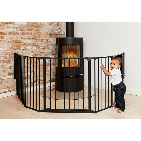 A baby standing in front of a fireplace with a black metal baby safety gate.