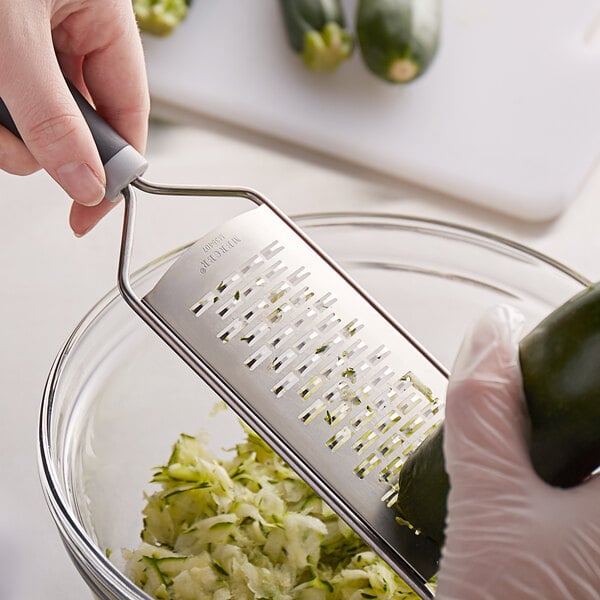 A gloved hand uses a MercerGrates ribbon grater with a black and grey handle to grate a green vegetable.