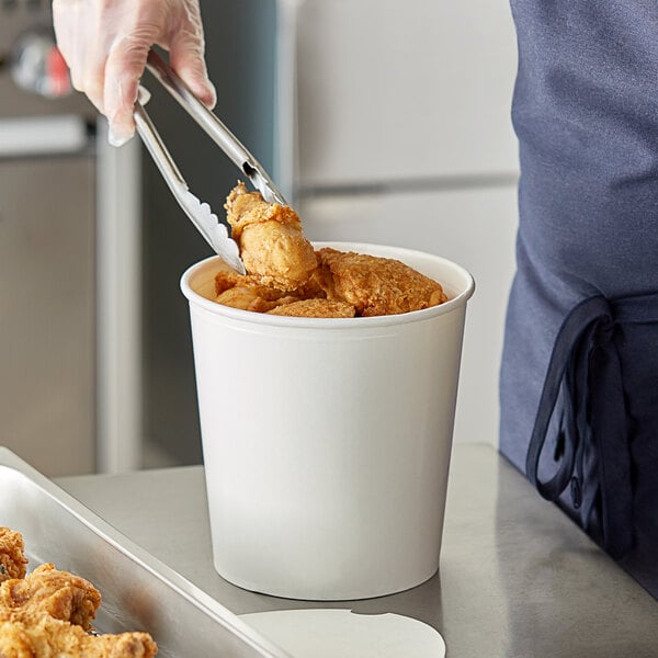 A person using metal tongs to serve chicken from a white food bucket.
